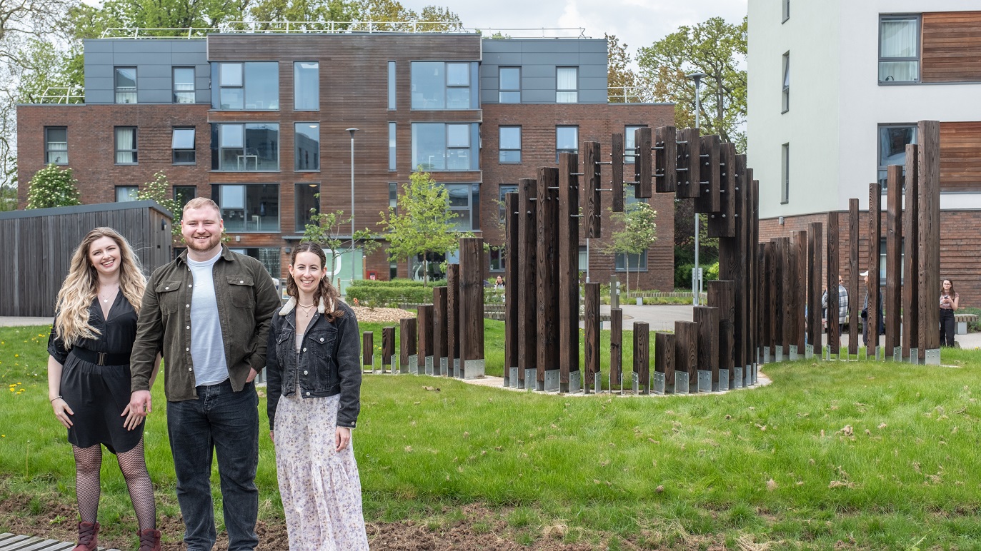 A wooden sculpture with multiple vertical panels of wood arising from a grassy mound in a wave like formation. There is a blank circular space in the middle of the sculpture. To the left three graduate students stand next to each other smiling at the camera.