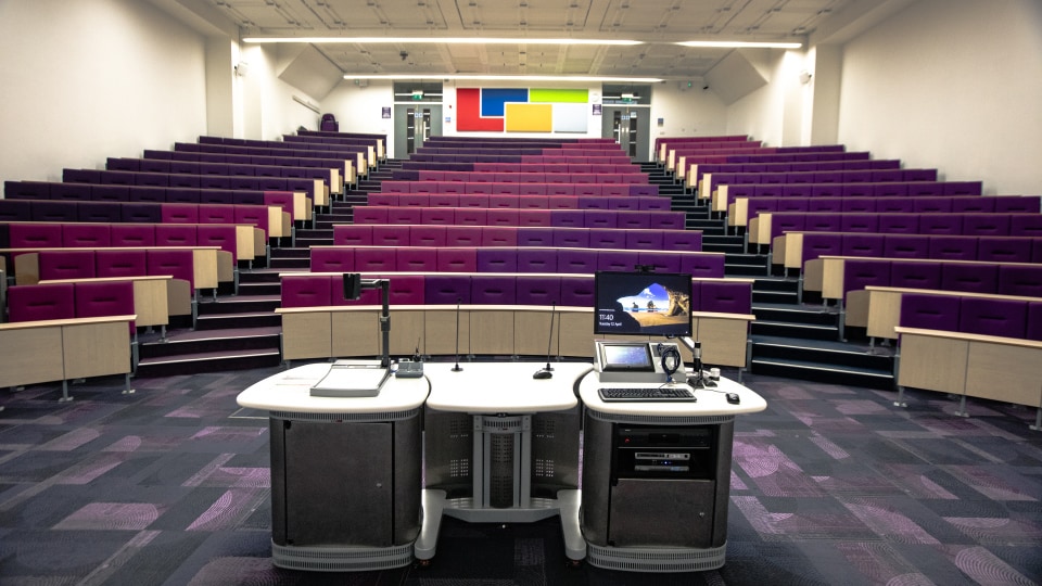 Wolfson T003 - Lecture theatre with tiered purple seating, central podium with computer, microphones, and projector. Colorful wall panels at back.