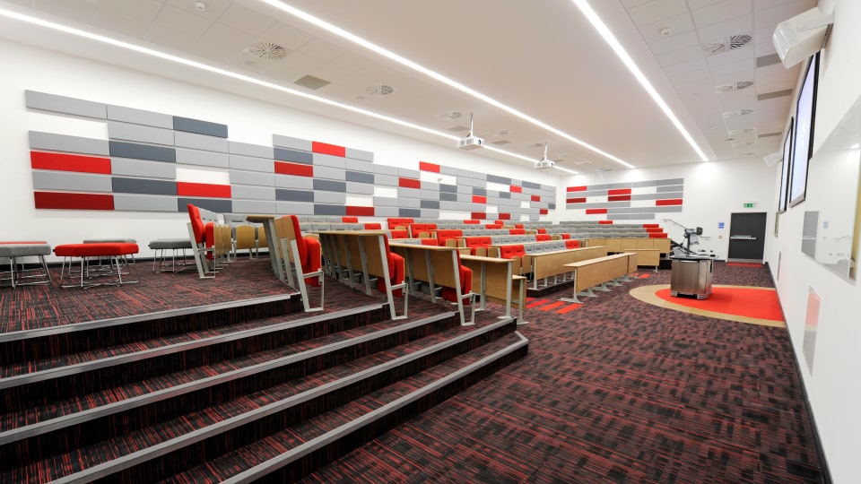 James France CC021 - Modern lecture hall with tiered seating, red and gray accents, and a podium. Bright lighting and wall panels.
