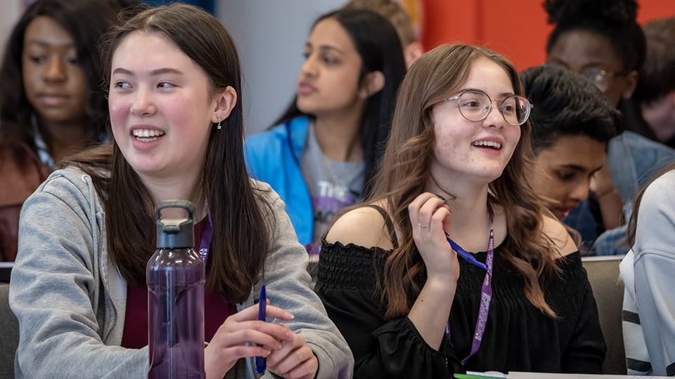 Two female students sitting side by side in a lecture theatre. More students sit behind them.