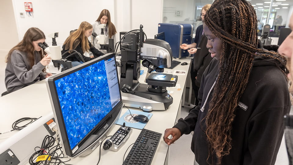 A female student standing at a desk looking at a computer screen. Students are in the background looking into microscopes