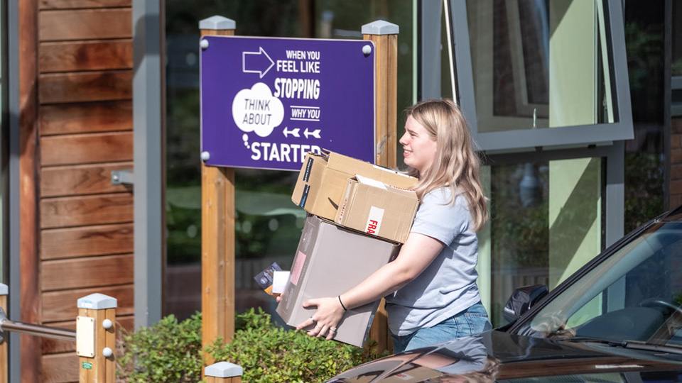A student carrying two boxes and walking outside a halls of residence on campus