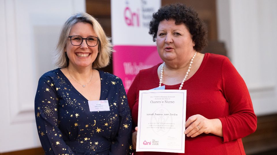 Two people standing together, the person on the right is holding a Queen’s Nurse certificate.
