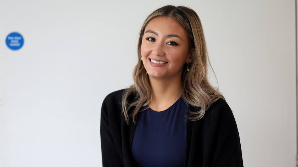 Headshot of PhD student Rhianna smiling at the camera, sat in front of a white wall, wearing a black cardi and a navy top.