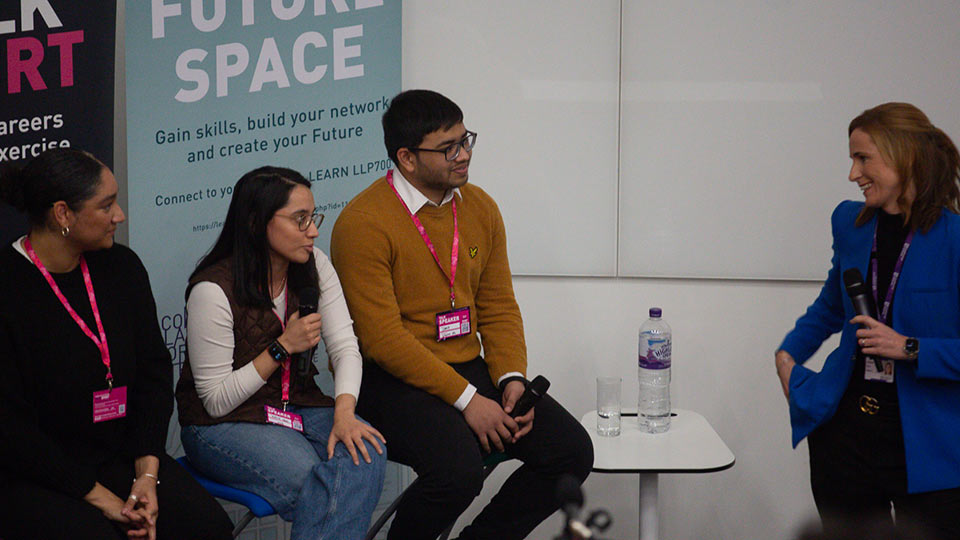Three people sat on chairs, one holding a microphones. Another person is standing, holding a microphones.