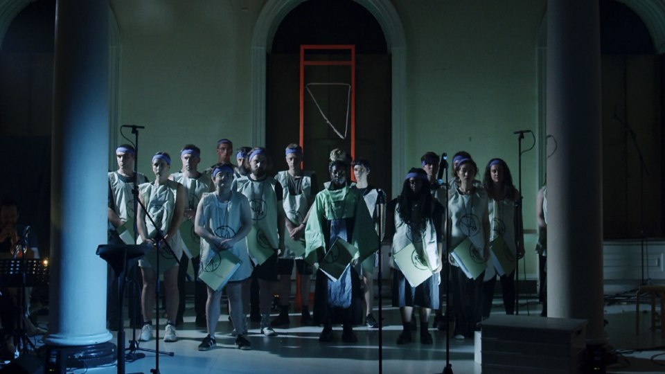 A choir standing together in a dark room behind microphones stands.