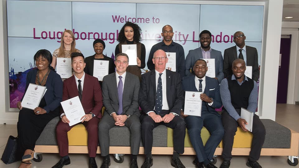 a group of students and professors posing for a photo at the London campus foyer presenting their certificates.