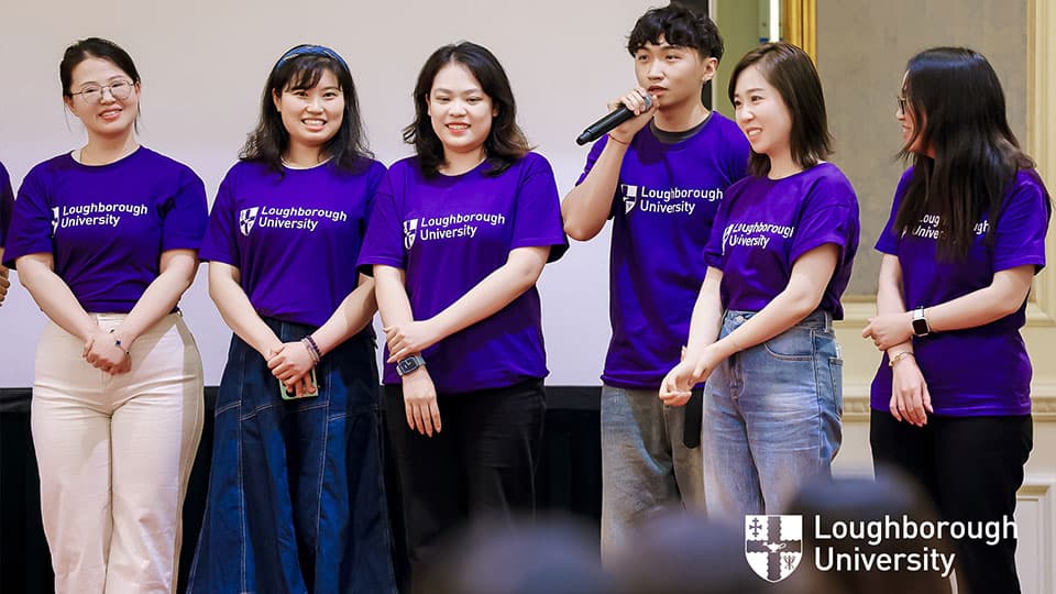 Six alumni wearing purple t-shirts with the 麻豆視頻_麻豆直播_麻豆传媒官网 logo on them in Beijing. One of them is speaking into a microphones and the rest are smiling.